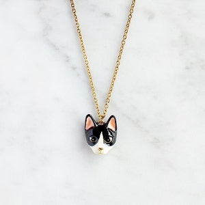 May include: A gold chain necklace with a black and white ceramic cat head pendant. The cat has pink ears and green eyes.
