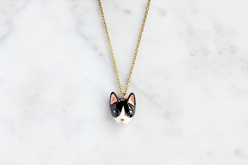 May include: A gold chain necklace with a black and white ceramic cat head pendant. The cat has pink ears and green eyes.