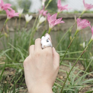May include: A white ceramic ring with a rabbit design. The ring is on a person's finger. The person's hand is reaching towards pink flowers.