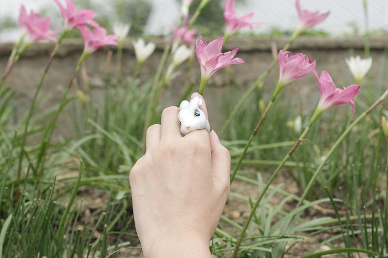 May include: A white ceramic ring with a rabbit design. The ring is on a person's finger. The person's hand is reaching towards pink flowers.