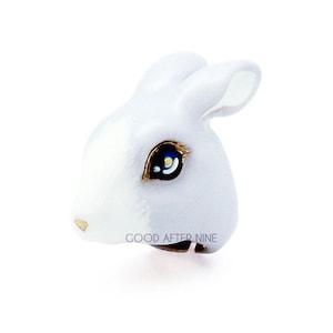 May include: White ceramic rabbit ring with a gold band and blue eyes. The ring says "GOOD AFTER NINE" on the band.