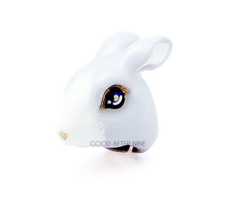 May include: White ceramic rabbit ring with a gold band and blue eyes. The ring says "GOOD AFTER NINE" on the band.