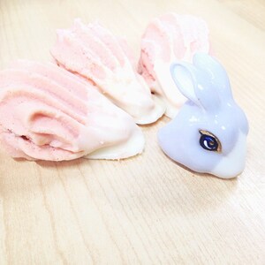 May include: Three pink and white meringue cookies and a purple ceramic bunny figurine on a light wood surface.