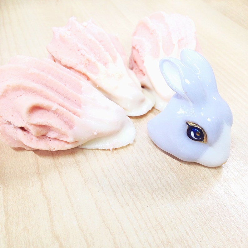 May include: Three pink and white meringue cookies and a purple ceramic bunny figurine on a light wood surface.