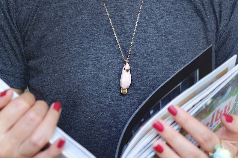 May include: A woman wearing a gray shirt with a gold chain necklace featuring a pink bird pendant. She is holding a stack of magazines.