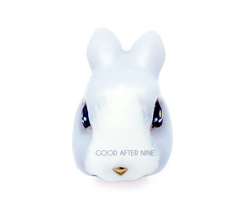 May include: A ceramic rabbit figurine with a white face and light blue ears. The rabbit has black eyes with white dots and a gold nose.