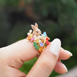 Puede incluir: Un anillo de oro con una figurita de conejo marrón rodeada de flores y hojas de colores.