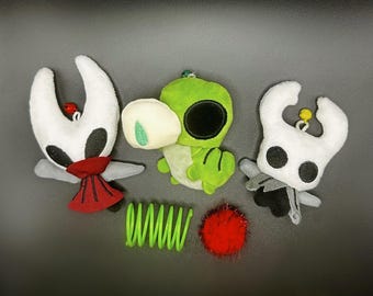 May include: Three plush toys: two white figures with black eyes and red accents, and a green creature with a large black eye. A green coil and a red pom-pom are also present. The toys are likely from a video game.