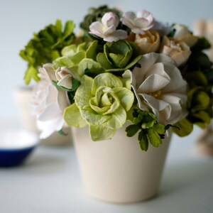 Puede incluir: Una maceta de cerámica blanca con un ramo de flores de papel blancas y verdes. Las flores están dispuestas en forma circular y están rodeadas de hojas verdes.