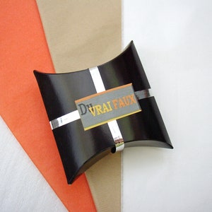 May include: A black pillow box with a silver ribbon and a white label with the text "DVRAI FAUX" in orange and gray letters.