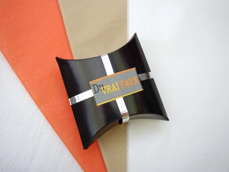 May include: A black pillow box with a silver ribbon and a white label with the text "DVRAI FAUX" in orange and gray letters.