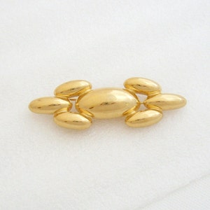 May include: A gold-toned brooch with a central oval shape and smaller oval shapes on either side. The brooch is made of smooth, shiny metal.