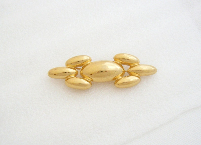 May include: A gold-toned brooch with a central oval shape and smaller oval shapes on either side. The brooch is made of smooth, shiny metal.
