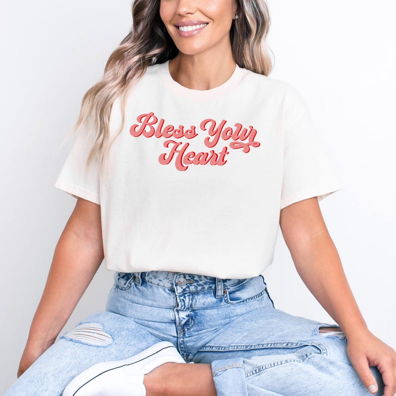 Puede incluir: Una camiseta blanca con un gr&aacute;fico rosa de estilo retro que dice "Bless Your Heart".