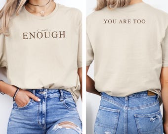 May include: A beige t-shirt with the words "I am ENOUGH" printed on the front in brown. The back of the shirt has the words "YOU ARE TOO" printed in brown. The shirt is paired with blue jeans.