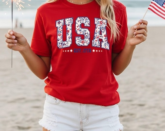 May include: A red t-shirt with the word "USA" in a floral design. The shirt also has the text "EST. 1776" and stars. The person is holding a sparkler and an American flag.