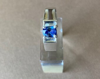 May include: A silver ring with a rectangular, bright blue gemstone. The ring has a modern design with a polished finish. The gemstone is set in a bezel setting, and the ring is displayed on a clear stand.