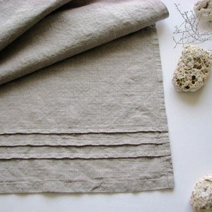 May include: A natural linen table runner in a subtle grey shade with a pleated design. The runner has a simple, elegant look and is perfect for adding a touch of rustic charm to any table setting.