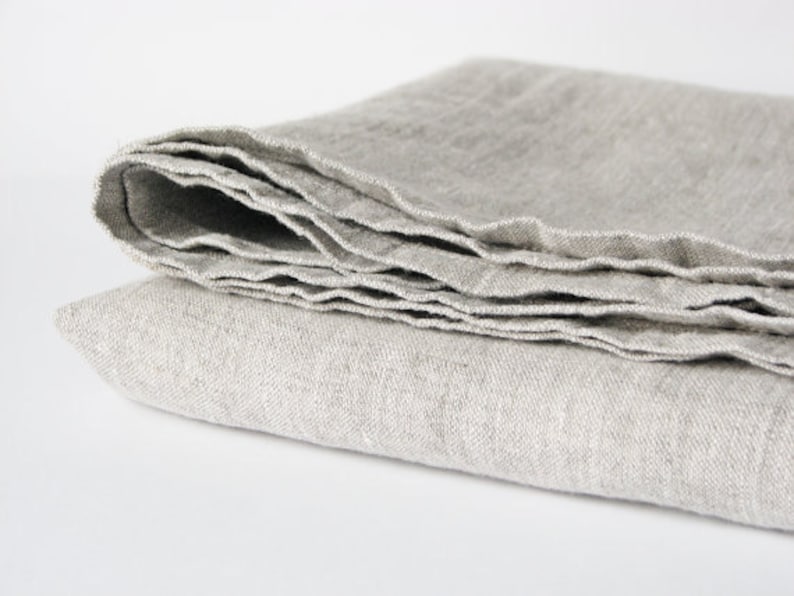 May include: A folded linen throw blanket in a light gray color. The blanket is made of natural fibers and has a soft, textured surface.
