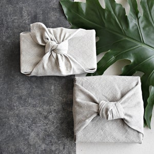 May include: Two gray fabric gift boxes tied with a knot. The boxes are on a gray textured surface with a green leaf in the background.
