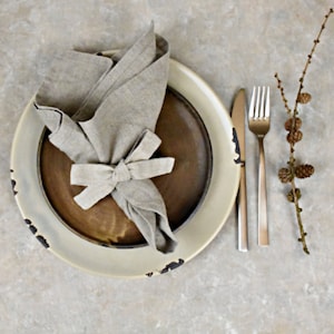 May include: A rustic, off-white dinner plate with a brown, glazed inner plate. A linen napkin is folded and tied into a bow on top of the inner plate. A silver fork and knife are placed to the right of the plate.