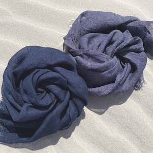 Op de afbeelding: Twee marineblauwe sjaals zijn op een zanderige ondergrond geplaatst. De sjaals zijn gemaakt van een zachte, lichtgewicht stof met een licht getextureerde uitstraling. De stof is gedrapeerd en gevouwen, wat een gevoel van beweging en textuur creëert.
