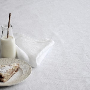 Op de afbeelding: Een plakje cake met poedersuiker erop, een glazen fles met een rietje en een witte doek servet op een wit oppervlak.