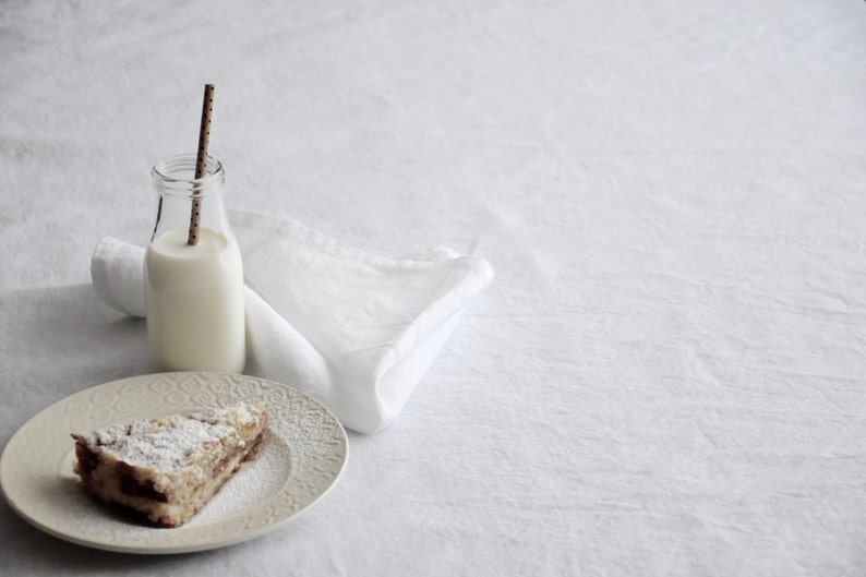 Op de afbeelding: Een plakje cake met poedersuiker erop, een glazen fles met een rietje en een witte doek servet op een wit oppervlak.