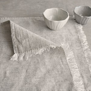 May include: A gray linen placemat with frayed edges. The placemat is folded in half and has a textured surface.