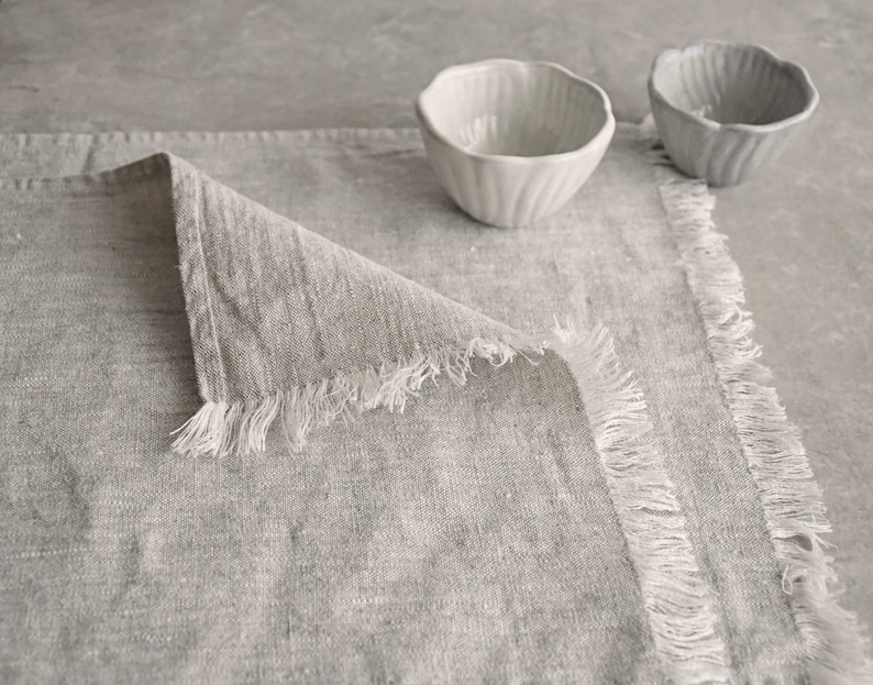 May include: A gray linen placemat with frayed edges. The placemat is folded in half and has a textured surface.