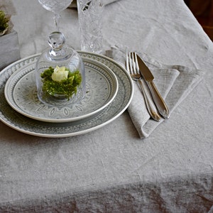 Op de afbeelding: Een grijs en wit gepatroneerd dinerbord met een kleiner grijs en wit gepatroneerd bord erop. Een glazen stolp met een witte bloem en groen mos erin staat op de borden. Een grijze linnen servet met een zilveren vork en mes ligt rechts van de borden.