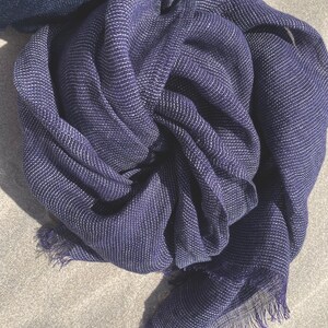 May include: A navy blue scarf with a textured weave and a fringe edge. The scarf has a subtle white thread running through the fabric, creating a striped pattern. The scarf is made of a lightweight material and is draped in soft folds.