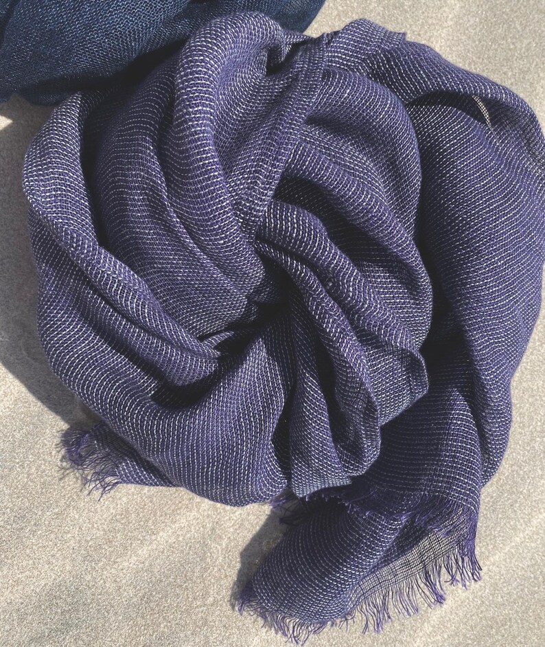 May include: A navy blue scarf with a textured weave and a fringe edge. The scarf has a subtle white thread running through the fabric, creating a striped pattern. The scarf is made of a lightweight material and is draped in soft folds.