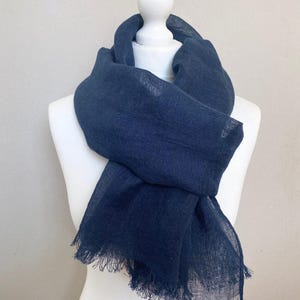 May include: A navy blue scarf with a fringed edge is draped over a white mannequin. The scarf is made of a lightweight fabric, and the texture is visible. The scarf is a solid color and appears to be a versatile accessory.
