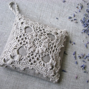 以下が含まれることがあります： 繊細な花柄のクロシェ編みで作られた正方形のラベンダーサシェ。サシェはナチュラルリネン生地で作られており、薄いベージュ色です。