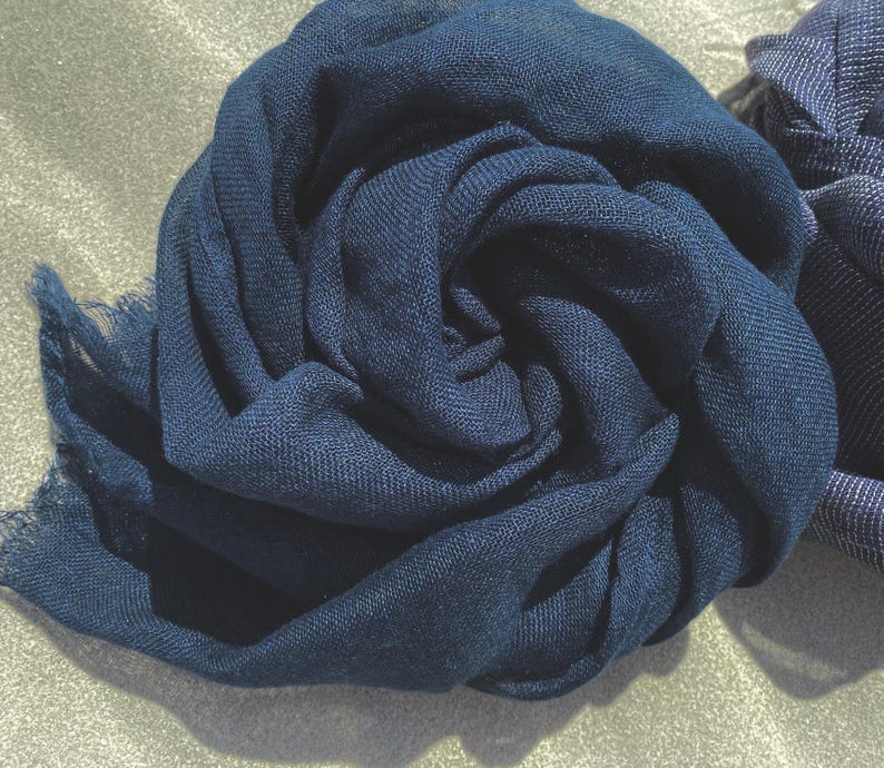 May include: A close-up of a navy blue scarf, artfully arranged in a spiral. The fabric has a soft texture and a fringed edge. Another scarf is partially visible on the right. The scarves are likely made of a lightweight material, perfect for adding a touch of elegance.