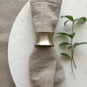 May include: A silver napkin ring with a slightly rounded shape is holding a folded linen napkin. The napkin is a light brown color and is placed on a white marble surface. A small green plant is in the background.