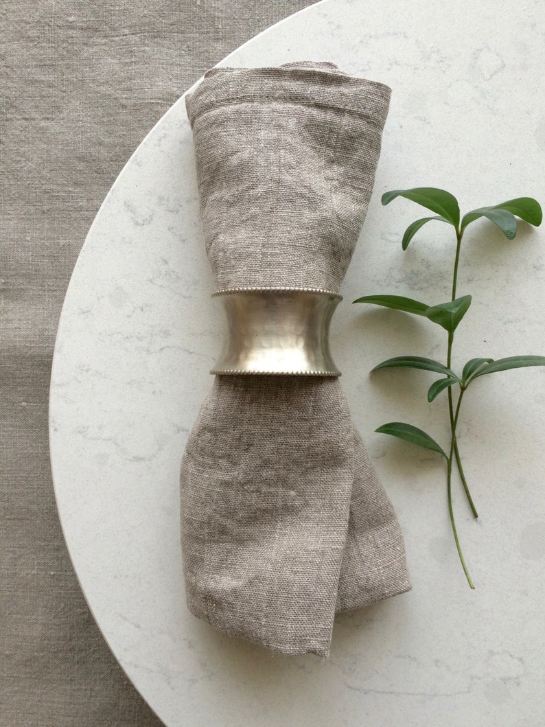 May include: A silver napkin ring with a slightly rounded shape is holding a folded linen napkin. The napkin is a light brown color and is placed on a white marble surface. A small green plant is in the background.