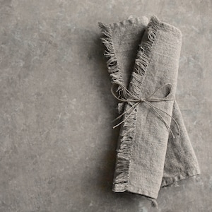 May include: Two linen napkins with frayed edges are tied together with twine. The napkins are a light brown color and have a rustic look.