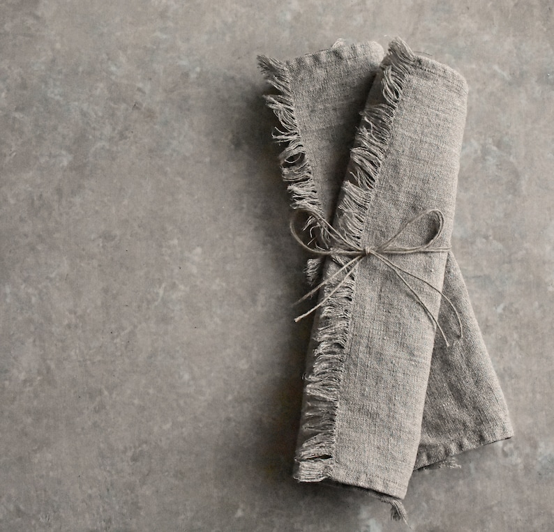 May include: Two linen napkins with frayed edges are tied together with twine. The napkins are a light brown color and have a rustic look.