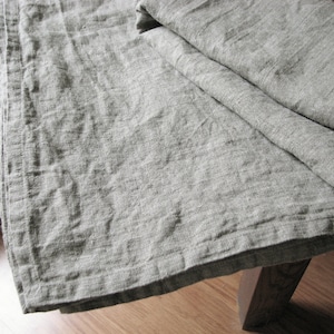 May include: A gray linen tablecloth with a textured weave. The tablecloth is folded in half and has a slightly wrinkled appearance.