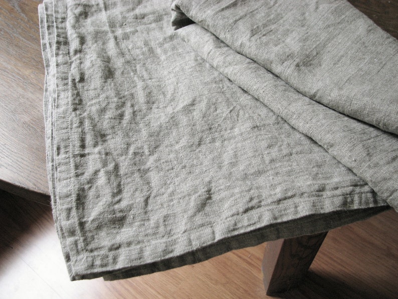 May include: A gray linen tablecloth with a textured weave. The tablecloth is folded in half and has a slightly wrinkled appearance.