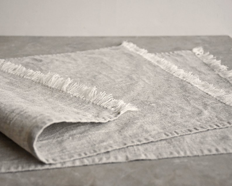 May include: A gray linen table runner with frayed edges. The runner is folded in half and has a textured surface.