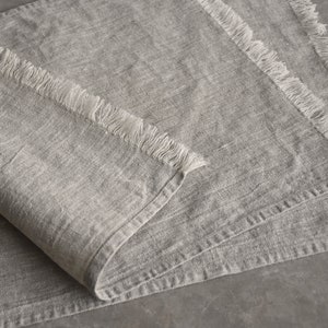May include: A gray linen table runner with a frayed edge. The runner is folded in half and has a soft, textured surface.