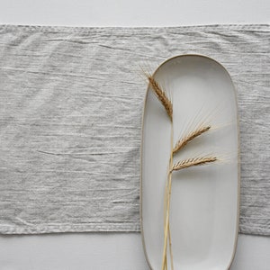 May include: A white oval ceramic platter with a gray linen placemat and a single sprig of dried wheat.