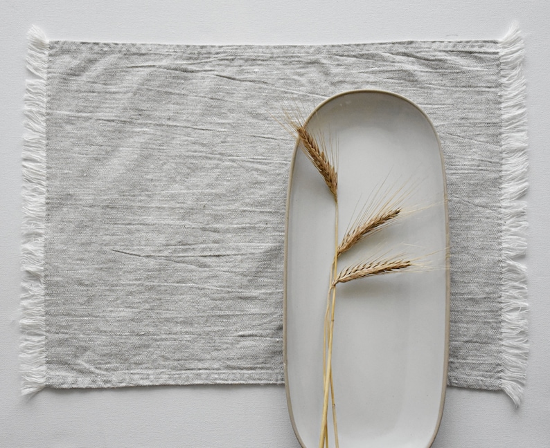 May include: A white oval ceramic platter with a gray linen placemat and a single sprig of dried wheat.