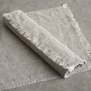 May include: A gray linen placemat with frayed edges. The placemat is folded in half and has a textured surface.