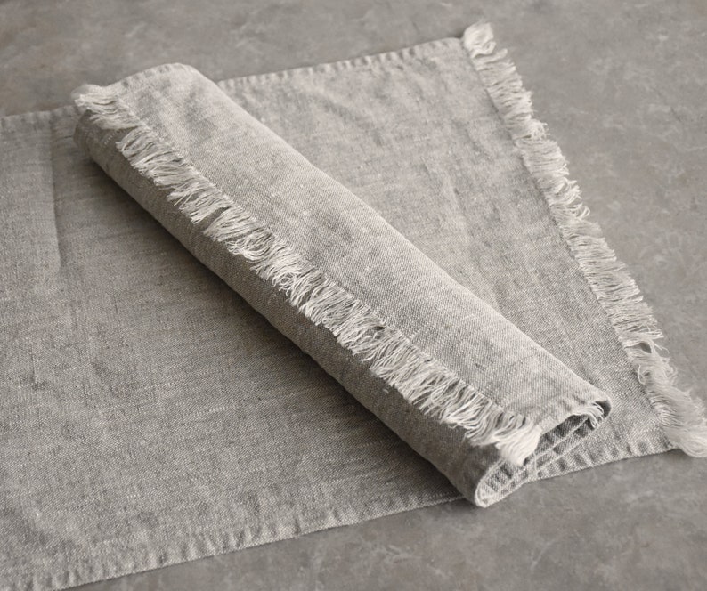 May include: A gray linen placemat with frayed edges. The placemat is folded in half and has a textured surface.