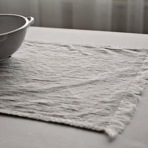 May include: A white linen placemat with a fringed edge. The placemat is textured and has a subtle sheen.