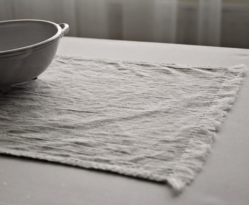 May include: A white linen placemat with a fringed edge. The placemat is textured and has a subtle sheen.